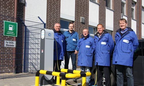 Fünf Männer an einer Elektro-Ladesäule vor einem Werksgebäude