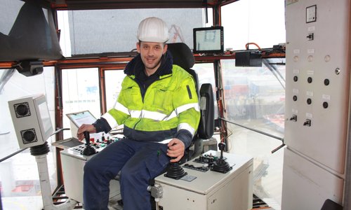 Mitarbeiter mit Warnjacke und Helm im Bedienstand eines Hafenkrans