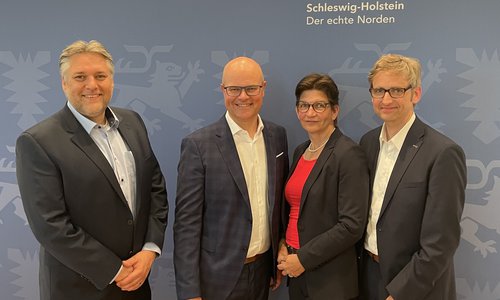 Vier Personen posieren vor einer Wand mit Schleswig-Holstein-Logo