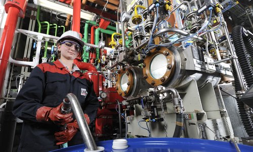 Auszubildende mit Schutzhelm an einer Chemieanlage mit blauem Fass