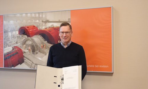 Mann hält einen geöffneten Aktenordner vor einem Foto einer Turbinenhalle