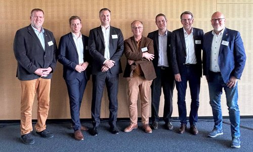 Gruppenfoto von sieben Männern in Anzügen vor einer Holzwand